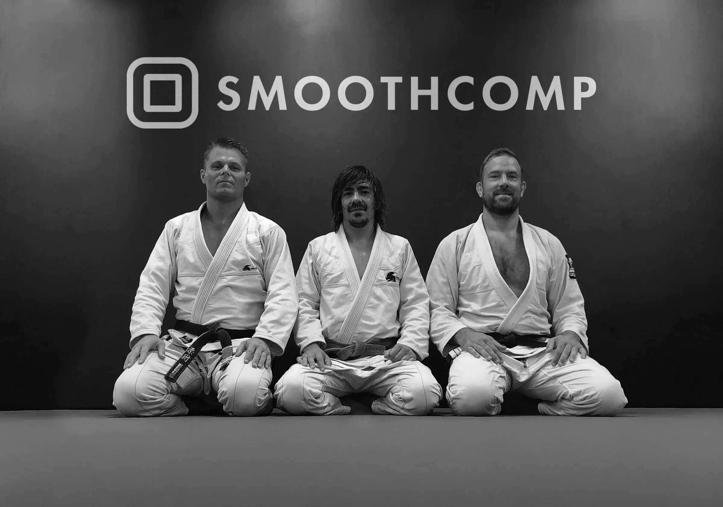 The three Smoothcomp co-founders in their gi, kneeling in front of the Smoothcomp logo